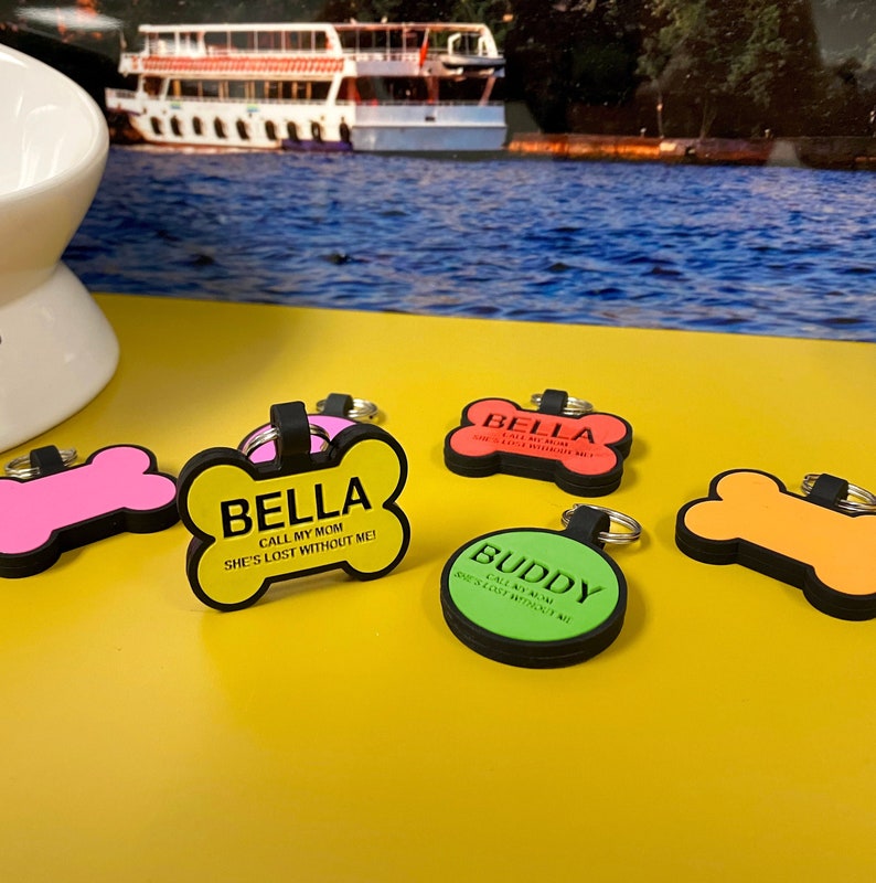 May include: Four colourful dog ID tags in the shape of bones and a circle. The tags have black text that reads "Bella, Call My Mom, She's Lost Without Me!" and "Buddy, Call My Mom, She's Lost Without Me!"