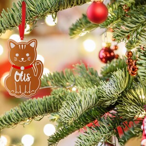 May include: A brown gingerbread cat ornament with a red ribbon and the name "Otis" written in white on the front. The ornament is hanging on a green Christmas tree.