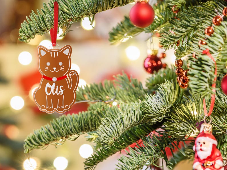 May include: A brown gingerbread cat ornament with a red ribbon and the name "Otis" written in white on the front. The ornament is hanging on a green Christmas tree.