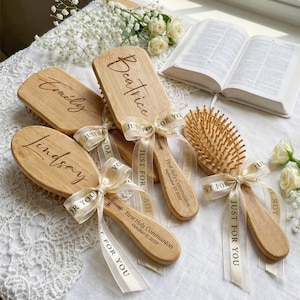 May include: Wooden hairbrushes with engraved names and dates, tied with cream-colored ribbons that read "Just For You." The brushes are personalized with names like "Emily," "Beatrice," and "Lindsay." The text "First Holy Communion" is also engraved.