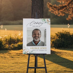 May include: A white sign on a black easel with the text "Celebrating the Life of George Lewis" and a photo of a smiling man. Floral accents frame the photo and text. The sign is displayed outdoors in a grassy area.