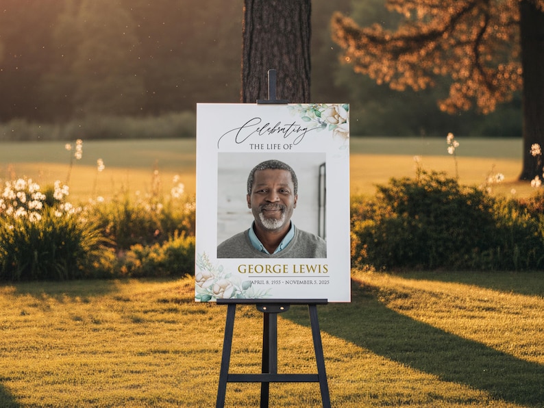 May include: A white sign on a black easel with the text "Celebrating the Life of George Lewis" and a photo of a smiling man. Floral accents frame the photo and text. The sign is displayed outdoors in a grassy area.