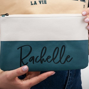 K&ouml;nnte beinhalten: Eine wei&szlig;e und t&uuml;rkisfarbene Rei&szlig;verschlusstasche mit dem Namen "Rachelle", der in Schwarz auf dem t&uuml;rkisfarbenen Teil gestickt ist.