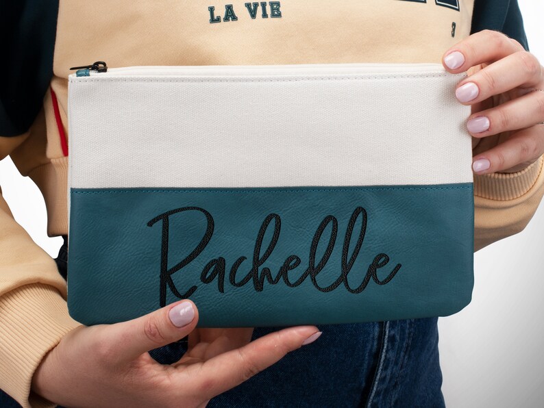 K&ouml;nnte beinhalten: Eine wei&szlig;e und t&uuml;rkisfarbene Rei&szlig;verschlusstasche mit dem Namen "Rachelle", der in Schwarz auf dem t&uuml;rkisfarbenen Teil gestickt ist.