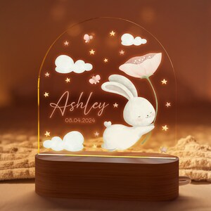 May include: A personalized night light featuring a white bunny holding a pink flower, with the name "Ashley" and the date "08.04.2024" displayed. The design includes clouds, stars, and butterflies, set on a wooden base.