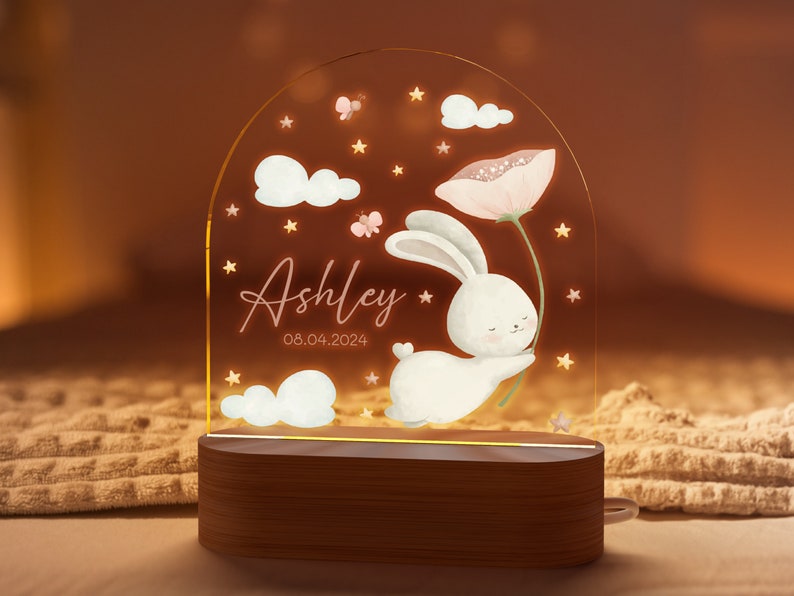 May include: A personalized night light featuring a white bunny holding a pink flower, with the name "Ashley" and the date "08.04.2024" displayed. The design includes clouds, stars, and butterflies, set on a wooden base.