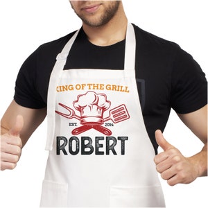 May include: White apron with a red chef hat, crossed grilling tools, and the text "King of the Grill Est. 2014 Robert".