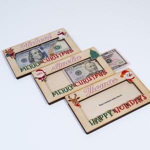 May include: Three wooden Christmas money holders with a red and green design. The holders have a slot for cash and a space for a personalized message. The names "Micheal", "Amelia", and "Thomas" are printed on the holders. The message "Merry Christmas" is printed on the top two holders and "Happy Holidays" is printed on the bottom holder.
