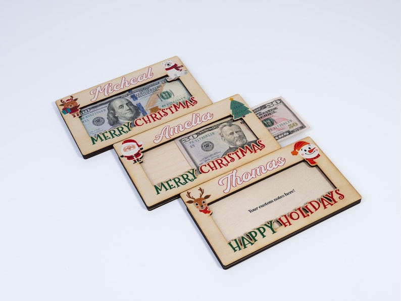 May include: Three wooden Christmas money holders with a red and green design. The holders have a slot for cash and a space for a personalized message. The names "Micheal", "Amelia", and "Thomas" are printed on the holders. The message "Merry Christmas" is printed on the top two holders and "Happy Holidays" is printed on the bottom holder.