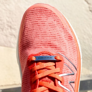 Puede incluir: Primer plano de una zapatilla de running naranja brillante con una etiqueta de metal plateado que dice "ROME 01.04.25".