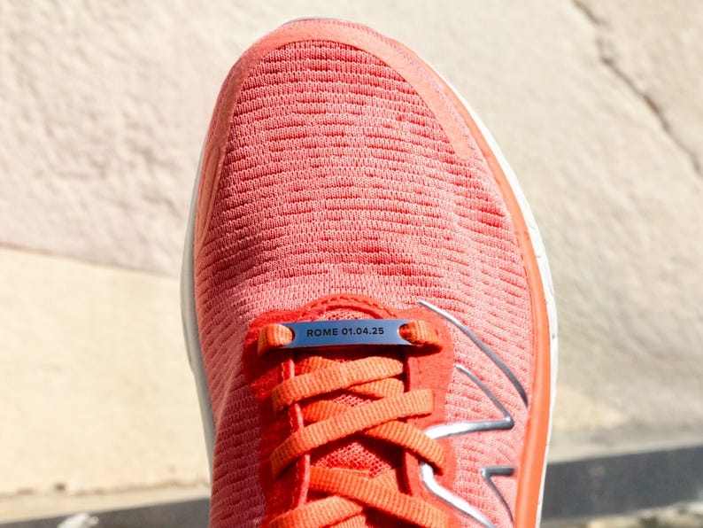 Puede incluir: Primer plano de una zapatilla de running naranja brillante con una etiqueta de metal plateado que dice "ROME 01.04.25".