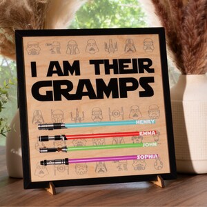 May include: Wooden framed sign with the text "I AM THEIR GRAMPS" and lightsaber designs. Below the text are four lightsabers with the names Henry, Emma, John, and Sophia. The sign is displayed on a wooden stand.