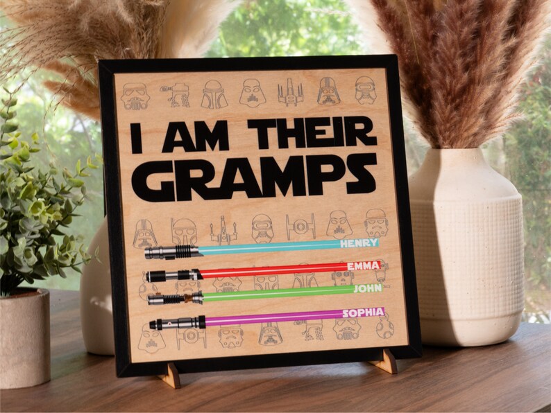May include: Wooden framed sign with the text "I AM THEIR GRAMPS" and lightsaber designs. Below the text are four lightsabers with the names Henry, Emma, John, and Sophia. The sign is displayed on a wooden stand.