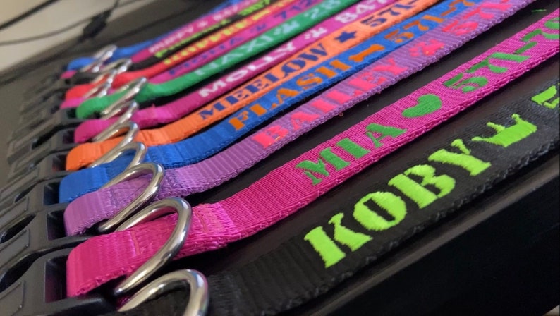 May include: A collection of colorful dog collars with names embroidered in various bright colors. The collars are made of a woven material and feature silver metal D-rings. Names include "Koby", "Mia", "Flash", and "MeeLow".