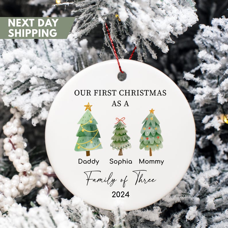 May include: White ceramic Christmas ornament with a watercolor illustration of three Christmas trees. The trees are labeled "Daddy", "Sophia", and "Mommy". The text "Our First Christmas as a Family of Three 2024" is printed on the ornament.