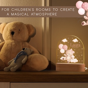 May include: A personalized night light with a teddy bear design, balloons, and the name "Laura" and date. The light sits on a wooden base. A large teddy bear and a smaller stuffed bear are also in the image.