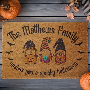 Pu&ograve; includere: Un tappetino per porta in fibra di cocco marrone con il testo "The Matthews Family wishes you a spooky Halloween" e tre gnomi dei cartoni animati che tengono zucche e caramelle.