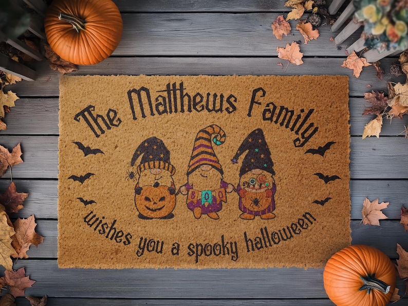Pu&ograve; includere: Un tappetino per porta in fibra di cocco marrone con il testo "The Matthews Family wishes you a spooky Halloween" e tre gnomi dei cartoni animati che tengono zucche e caramelle.