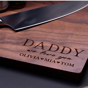 May include: A wooden cutting board with the text "Daddy we love you Olivia &hearts; Mia &hearts; Tom" engraved on it. The board is brown and has a natural wood grain.