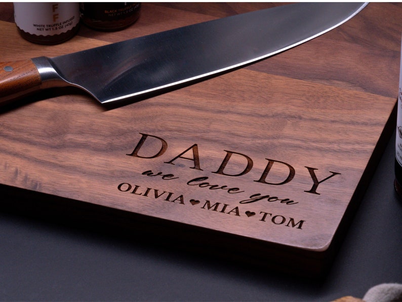 May include: A wooden cutting board with the text "Daddy we love you Olivia &hearts; Mia &hearts; Tom" engraved on it. The board is brown and has a natural wood grain.