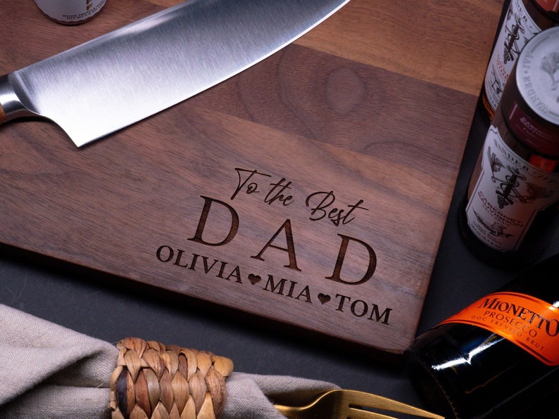 May include: A wooden cutting board with the text "To the Best Dad Olivia Mia Tom" engraved on it. The board is brown and has a natural wood grain. There is a knife and a bottle of wine in the background.
