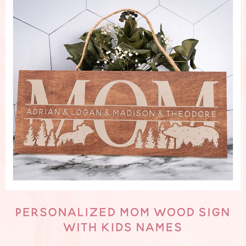 May include: Personalized wooden sign with the word "MOM" in large letters and the names "Adrian & Logan & Madison & Theodore" in a smaller font below. The sign is decorated with a woodland scene featuring trees and bears.
