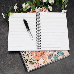 May include: An open spiral bound notebook with white lined pages and a black pen lying on top. The notebook is on top of a floral patterned notebook with pink and green flowers.