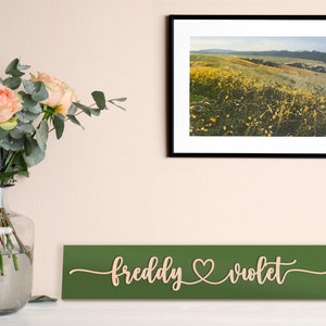 May include: A green wooden sign with the names "freddy" and "violet" written in cursive with a heart symbol between them. The sign is hanging on a wall with a framed photograph of a field of yellow wildflowers above it.