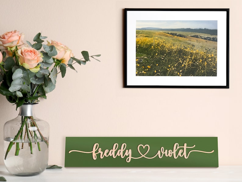 May include: A green wooden sign with the names "freddy" and "violet" written in cursive with a heart symbol between them. The sign is hanging on a wall with a framed photograph of a field of yellow wildflowers above it.