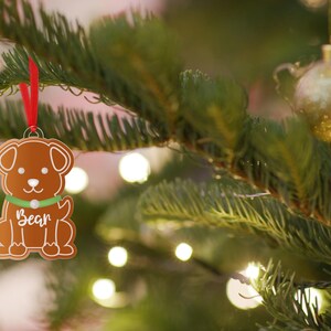 May include: A brown gingerbread dog ornament with a green collar and the name "Bear" written on it. The ornament is hanging on a green Christmas tree with white lights.