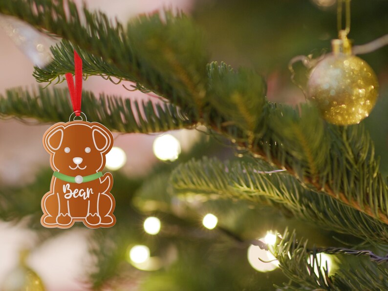 May include: A brown gingerbread dog ornament with a green collar and the name "Bear" written on it. The ornament is hanging on a green Christmas tree with white lights.