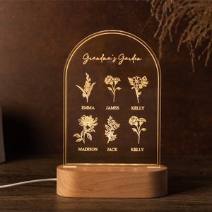 May include: A personalized LED night light with a wooden base. The clear acrylic panel features the text "Grandma's Garden" and floral illustrations with names below. The light is illuminated, showcasing the engraved design. The overall design is a warm, inviting home decor item.