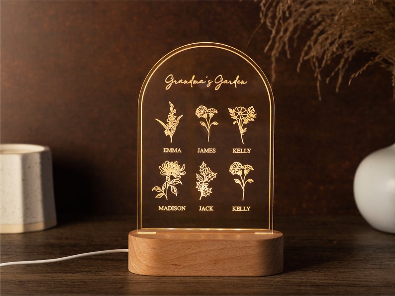 May include: A personalized LED night light with a wooden base. The clear acrylic panel features the text "Grandma's Garden" and floral illustrations with names below. The light is illuminated, showcasing the engraved design. The overall design is a warm, inviting home decor item.