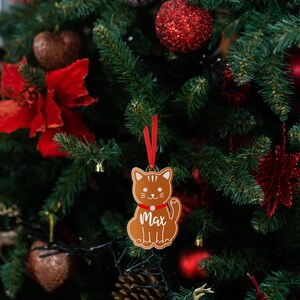 May include: A gingerbread-style cat ornament with a red ribbon and the name "Max" hanging on a Christmas tree with red and green decorations.