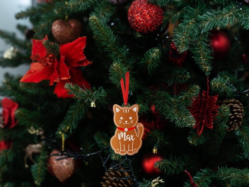 May include: A gingerbread-style cat ornament with a red ribbon and the name "Max" hanging on a Christmas tree with red and green decorations.