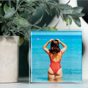 K&ouml;nnte beinhalten: Ein Glas-Bilderrahmen mit einem Bild einer Person in einem roten Badeanzug, die im Meer steht. Das Meer ist blau und der Himmel ist klar.