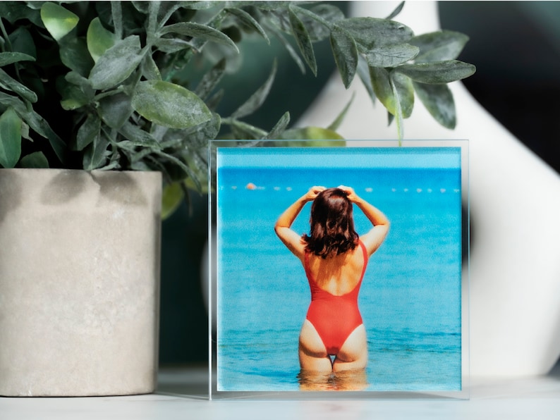 K&ouml;nnte beinhalten: Ein Glas-Bilderrahmen mit einem Bild einer Person in einem roten Badeanzug, die im Meer steht. Das Meer ist blau und der Himmel ist klar.