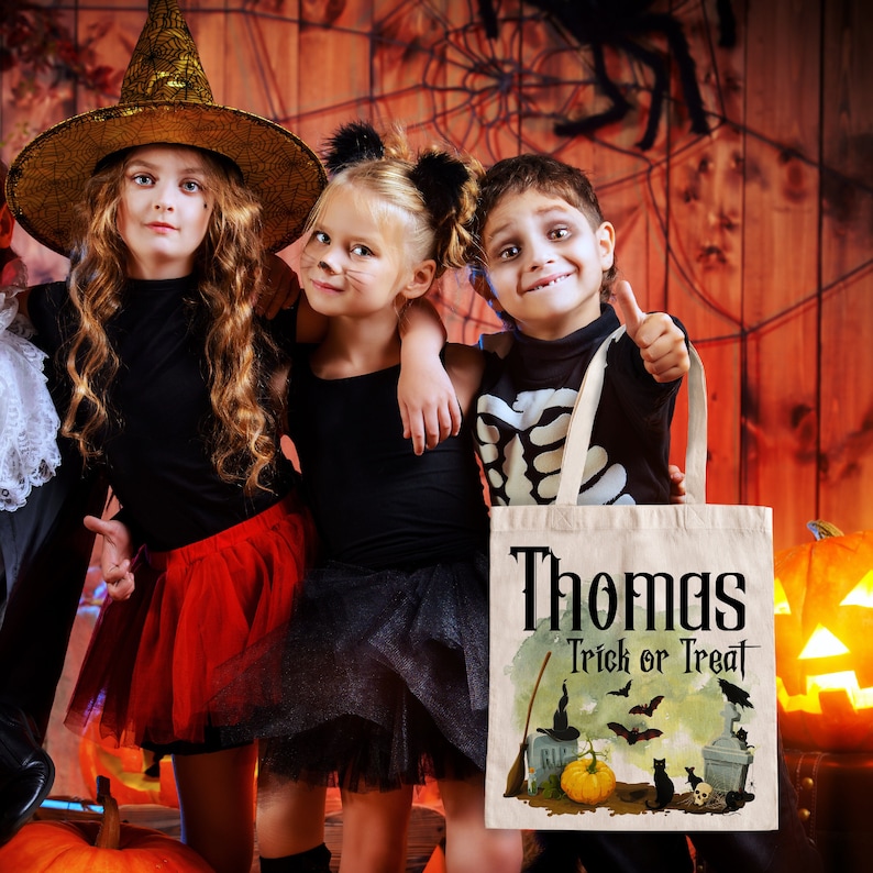 May include: Three children dressed in Halloween costumes, a witch, a cat, and a skeleton, holding a white canvas trick or treat bag with the name "Thomas" and the text "Trick or Treat" printed on it. The bag features a Halloween scene with a witch's hat, bats, a pumpkin, and a black cat.