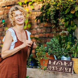 May include: A brown wooden sign with the word "MOM" in large white letters and the text "Samuel & Cooper & Aubrey" in smaller white letters below. The sign is decorated with white flowers and green leaves.