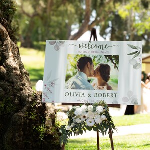 May include: A wedding welcome sign with the words "welcome to our beginning" and a photo of a couple kissing. The sign also includes the names Olivia & Robert and the date August 18, 2024. The sign is decorated with floral accents.