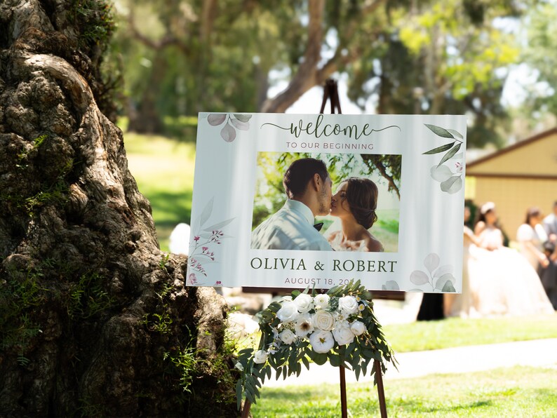 May include: A wedding welcome sign with the words "welcome to our beginning" and a photo of a couple kissing. The sign also includes the names Olivia & Robert and the date August 18, 2024. The sign is decorated with floral accents.
