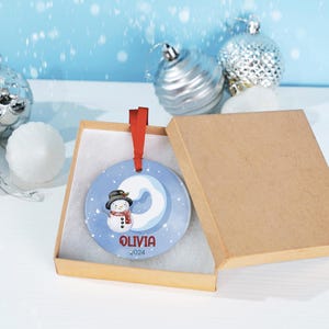 May include: A blue and white ceramic Christmas ornament with a snowman and the name "Olivia" and the year "2024" in red lettering. The ornament is in a brown gift box with a white interior.
