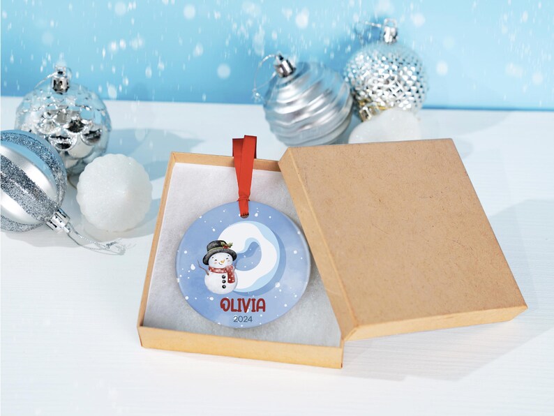 May include: A blue and white ceramic Christmas ornament with a snowman and the name "Olivia" and the year "2024" in red lettering. The ornament is in a brown gift box with a white interior.