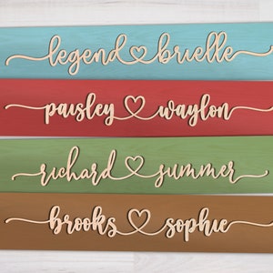May include: Four wooden signs with different colored backgrounds and the names of couples written in cursive with a heart symbol between each name. The names are: Legend & Brielle, Paisley & Waylon, Richard & Summer, Brooks & Sophie.