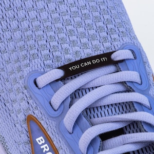 Puede incluir: Un par de zapatillas de running azul claro con un cierre de encaje negro que dice "YOU CAN DO IT!" Las zapatillas tienen un logotipo marr&oacute;n en el lateral.