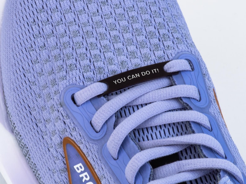Puede incluir: Un par de zapatillas de running azul claro con un cierre de encaje negro que dice "YOU CAN DO IT!" Las zapatillas tienen un logotipo marr&oacute;n en el lateral.