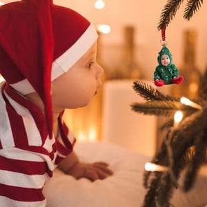Pu&ograve; includere: Un bambino con un vestito a righe rosse e bianche e un cappello da elfo rosso guarda un ornamento per l'albero di Natale. L'ornamento &egrave; un bambino piccolo con un costume da albero di Natale verde.