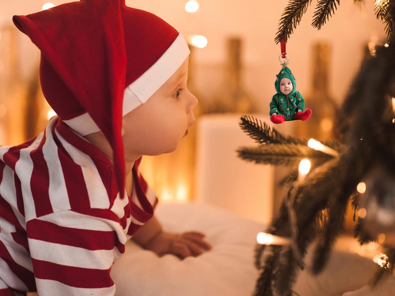 Pu&ograve; includere: Un bambino con un vestito a righe rosse e bianche e un cappello da elfo rosso guarda un ornamento per l'albero di Natale. L'ornamento &egrave; un bambino piccolo con un costume da albero di Natale verde.