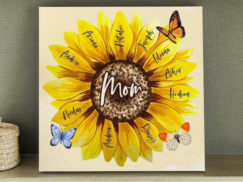 May include: A square canvas print featuring a watercolor sunflower design. The center reads "Mom" in white script. Each petal displays a name in black script. Butterflies in shades of orange, blue, and white are scattered around the flower.