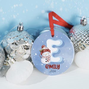 May include: A blue and white ceramic Christmas ornament with a snowman and the letter E. The ornament is personalized with the name "emily" and the year "2024".
